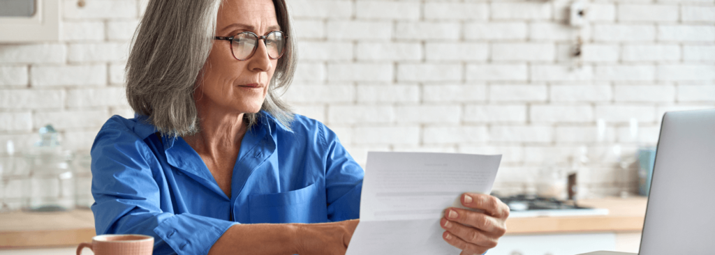 Adult senior woman working at home at laptop holding document calculating taxes